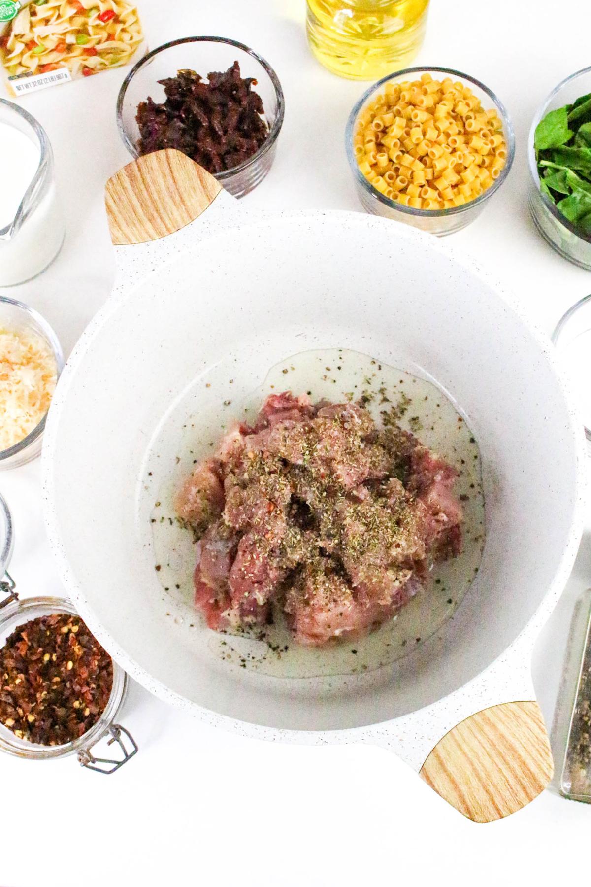 Raw seasoned meat in a white pot, surrounded by bowls of dry pasta, spinach, cheese, oil, spices, and chopped ingredients on a white surface.