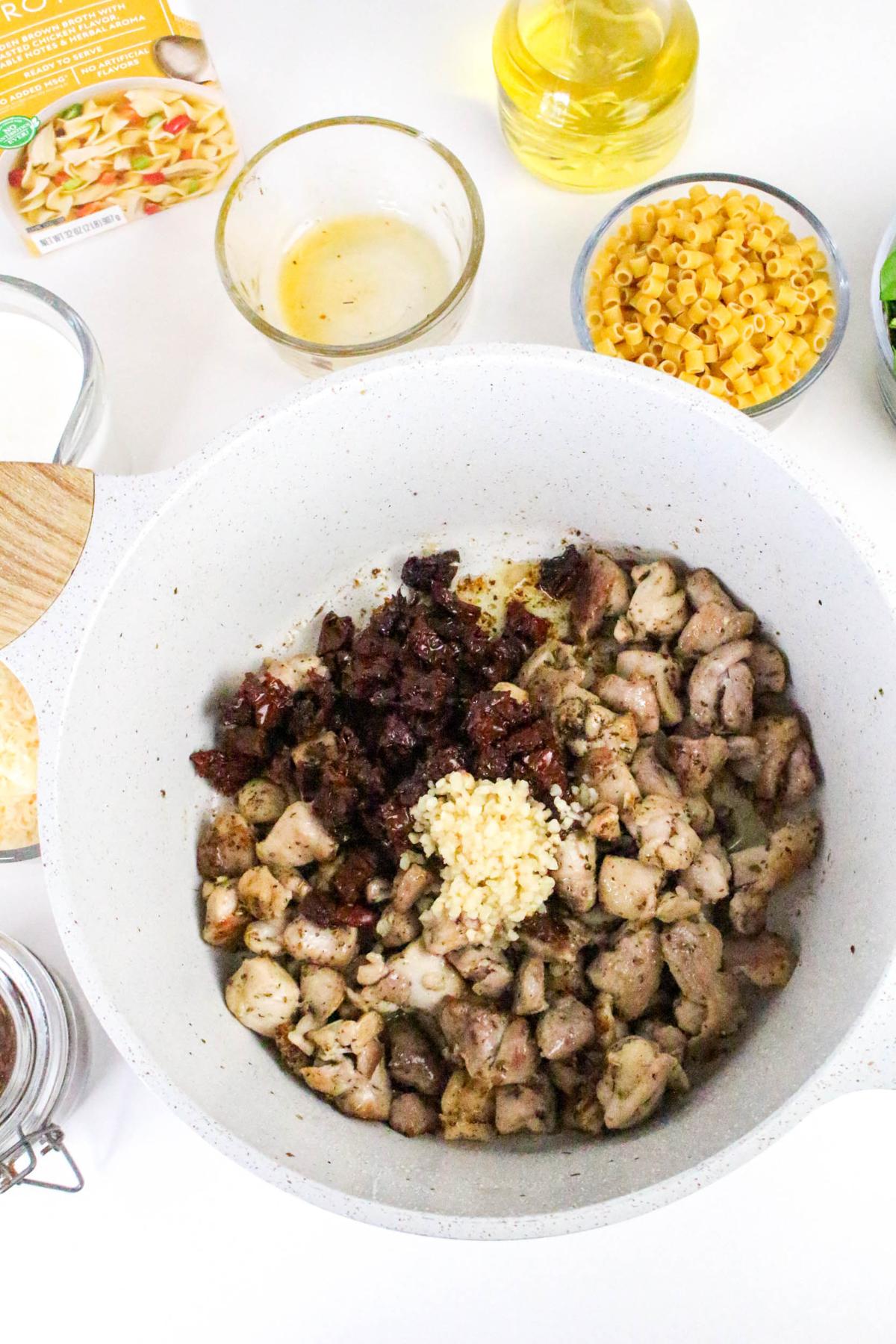 A pot containing cooked chicken pieces, minced garlic, and chopped sun-dried tomatoes is surrounded by bowls of dry pasta, oil, milk, and seasonings on a white surface.