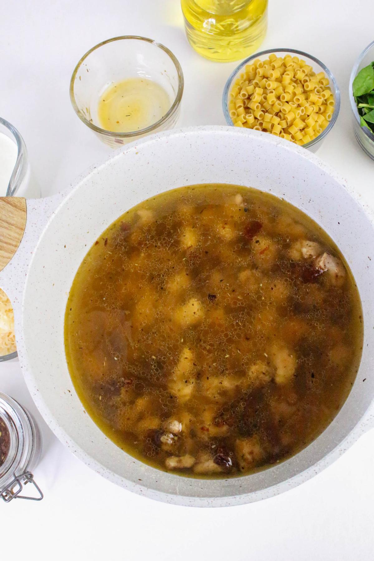 A pot of soup with chunks of meat, surrounded by small bowls containing uncooked pasta, greens, oil, and other ingredients on a white surface.