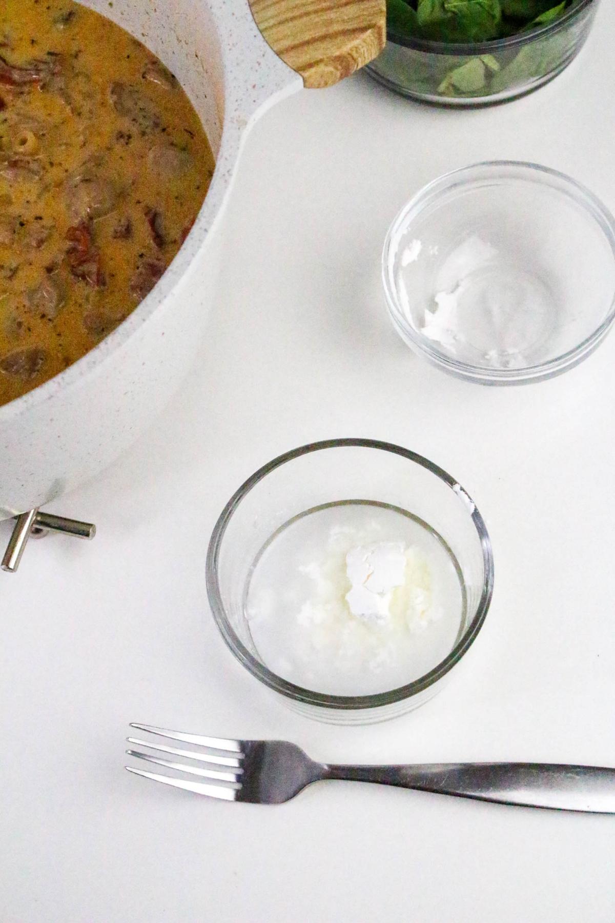 A pot of creamy soup sits next to small bowls containing white powder and liquid, with a fork placed on the white surface nearby.