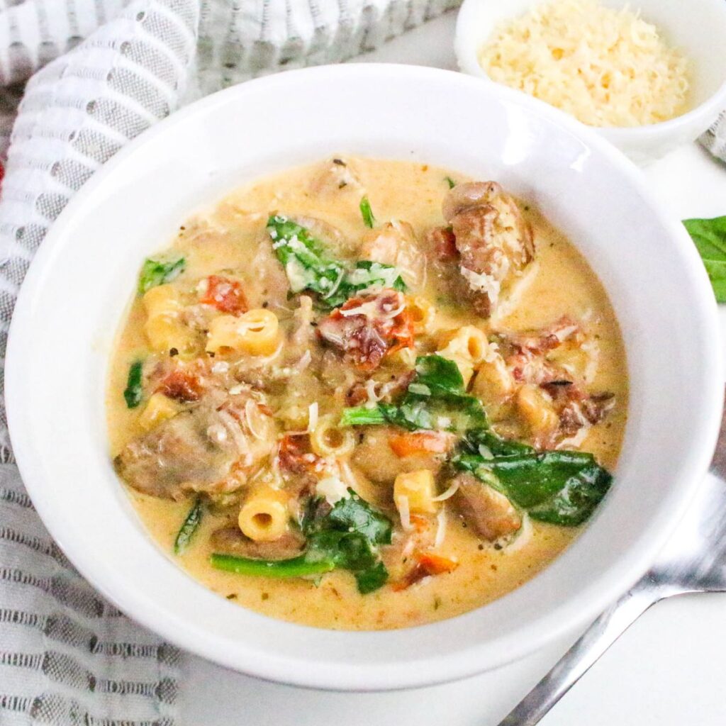 A white bowl filled with creamy soup containing pieces of chicken, pasta, spinach, and tomatoes, with a spoon nearby and a small dish of grated cheese in the background.