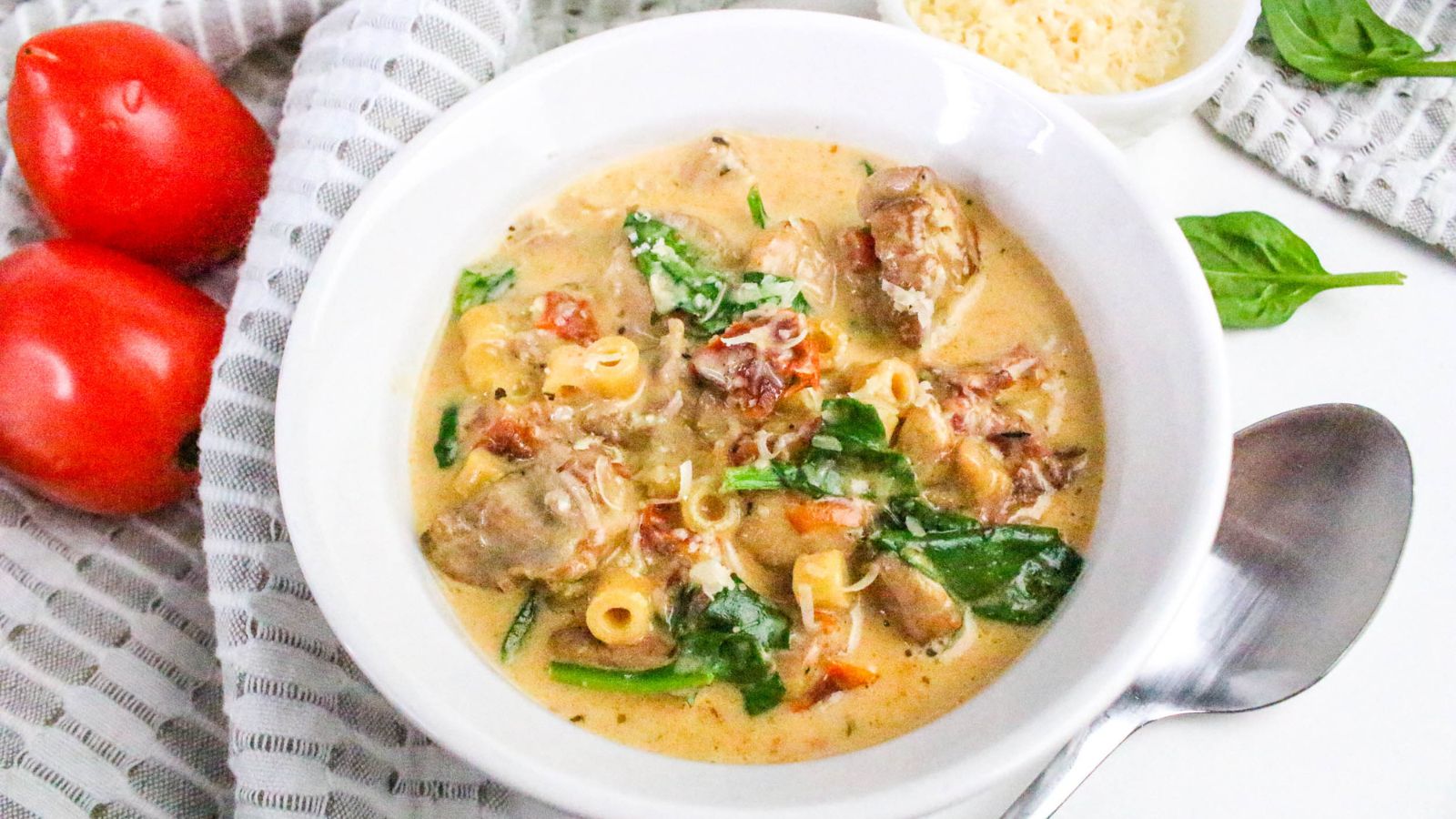 A bowl of creamy soup with pasta, spinach, tomatoes, and chicken pieces, placed next to two tomatoes, a spoon, and a small bowl of grated cheese.