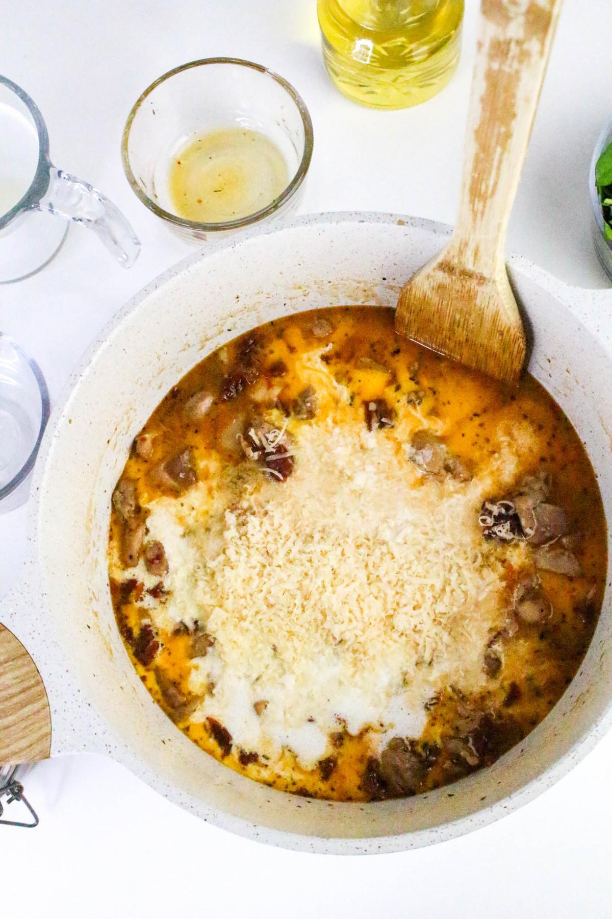 A pot of creamy soup with sausage, bacon, and grated cheese being stirred with a wooden spoon. Various bowls with ingredients are nearby on a white surface.