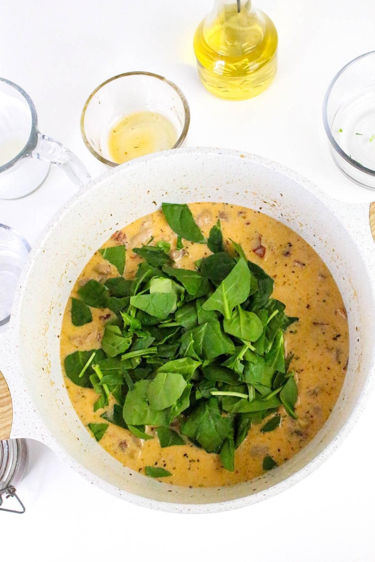 A pot of creamy soup with spinach leaves on top, surrounded by clear bowls and a bottle of olive oil on a white surface.
