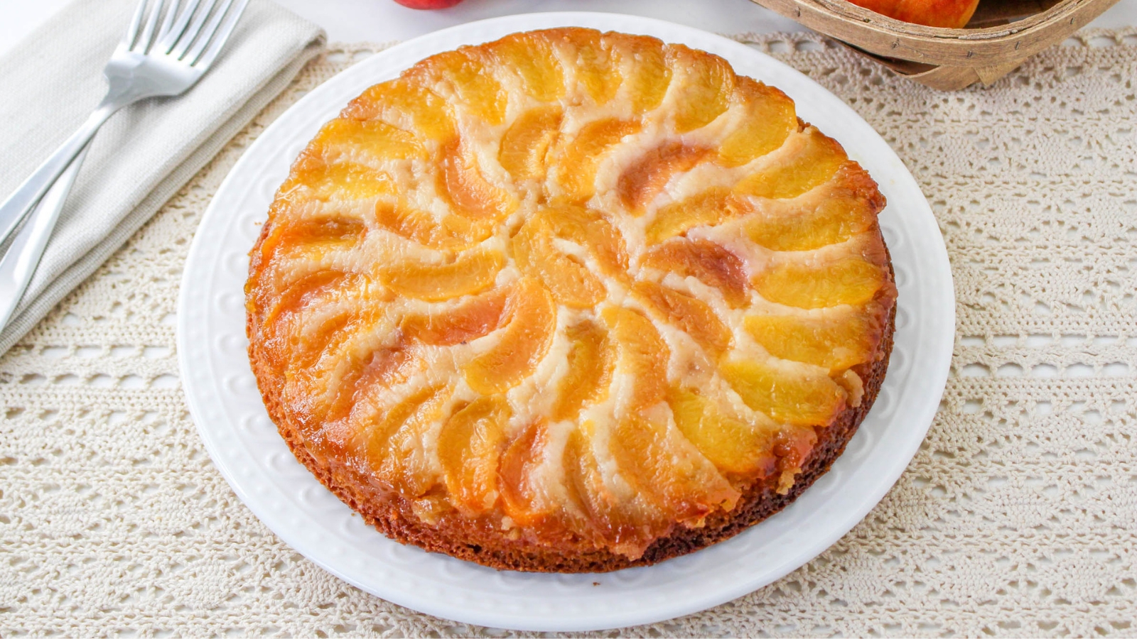A round peach upside-down cake on a white plate, displaying sliced peaches caramelized on top, set on a lace tablecloth with two forks on a napkin nearby.