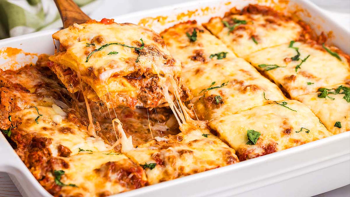 A baked ravioli lasagna in a white dish with cheese and basil on top. A piece is being lifted, showing melted cheese stretching from the dish.