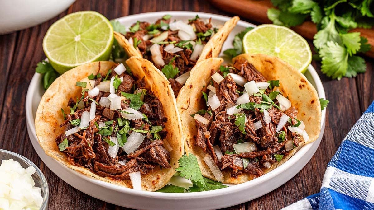 Three beef tacos with chopped onions and cilantro, served with lime halves, on a plate.