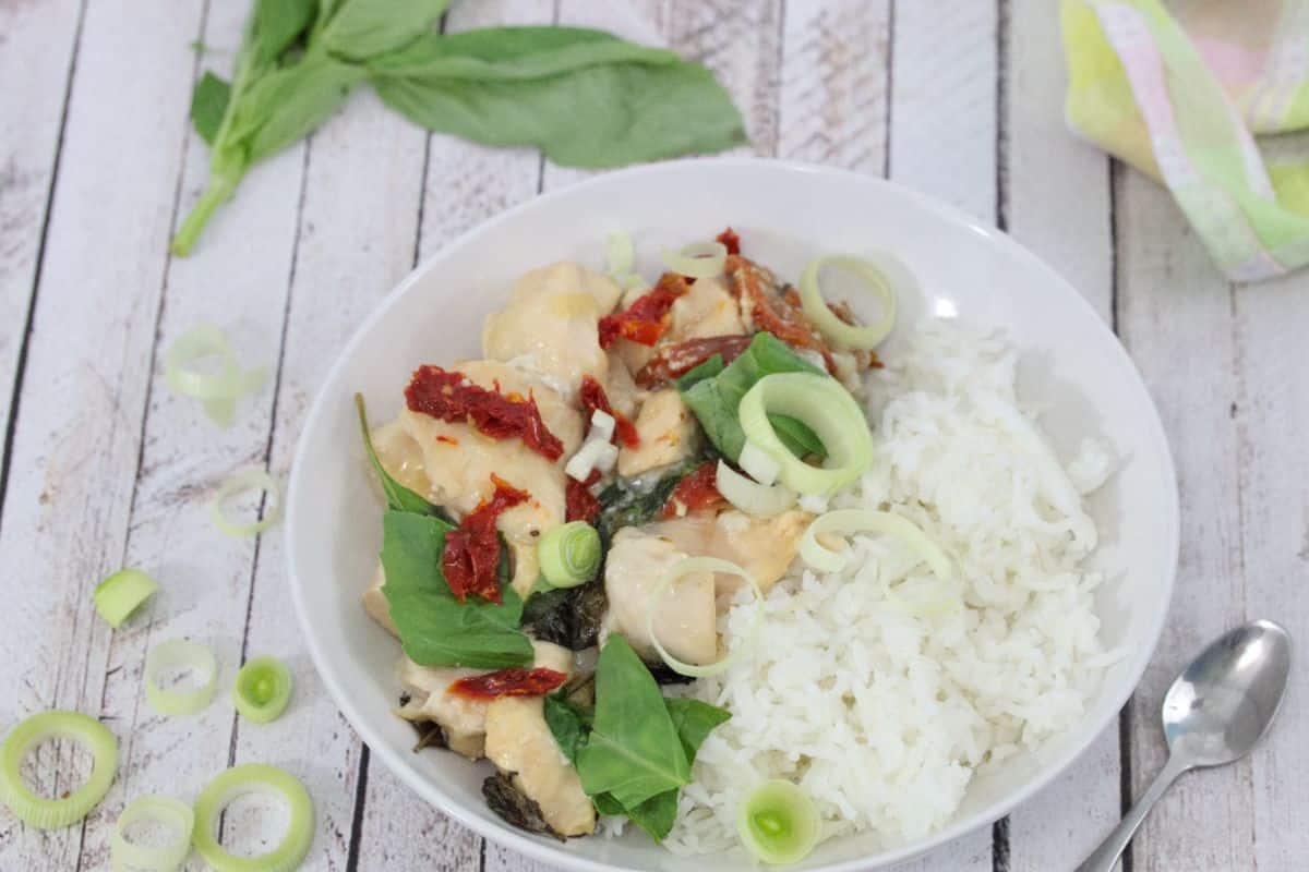 White bowl with tuscan chicken, rice, leeks and sundried tomato in it with fresh basil beside it.