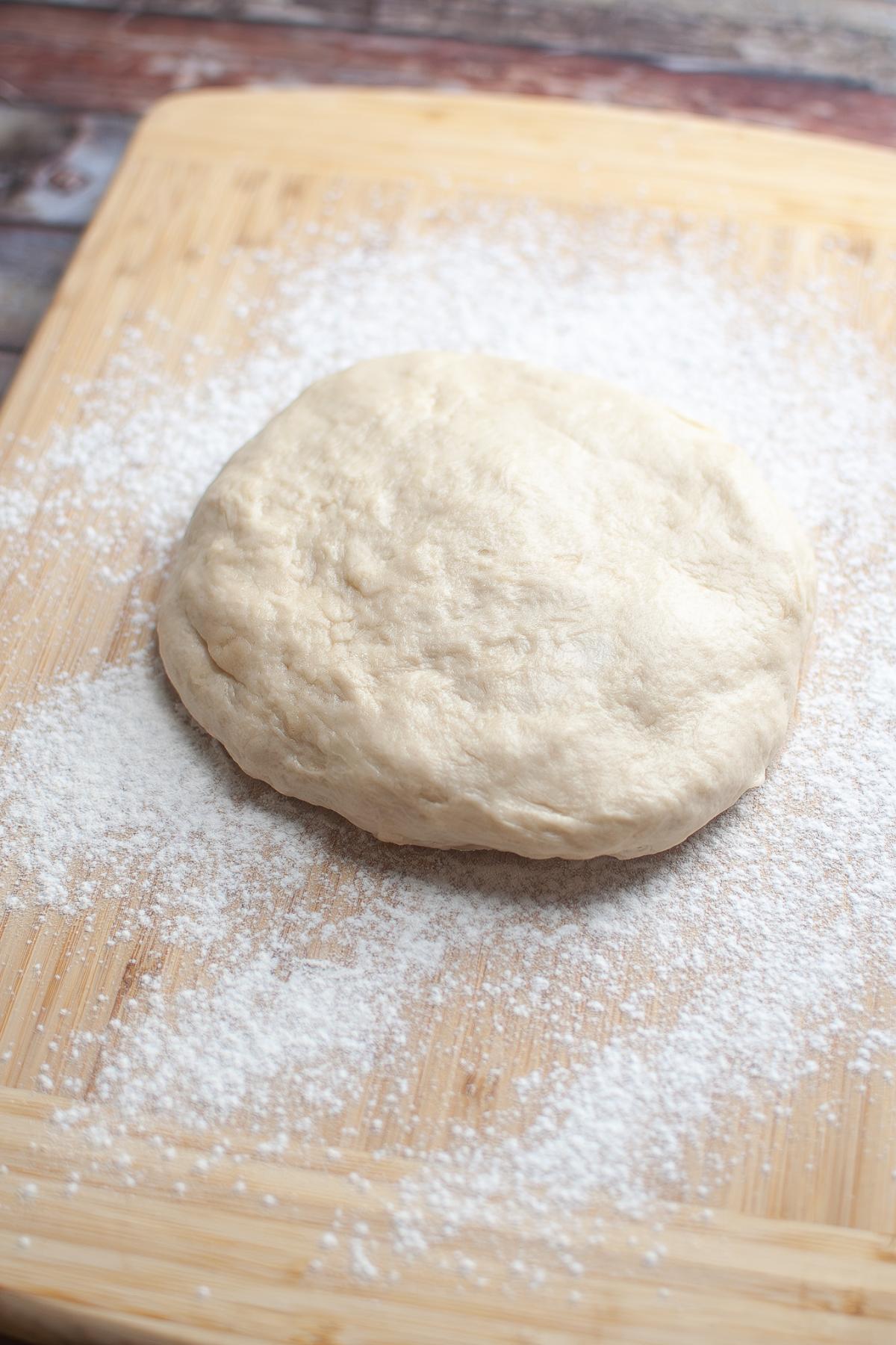 A round ball of raw pizza dough rests on a floured wooden cutting board.