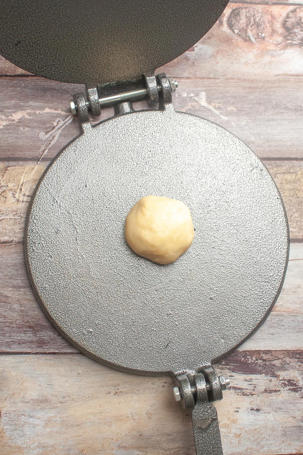 A ball of dough sits in the center of an open metal tortilla press on a wooden surface.