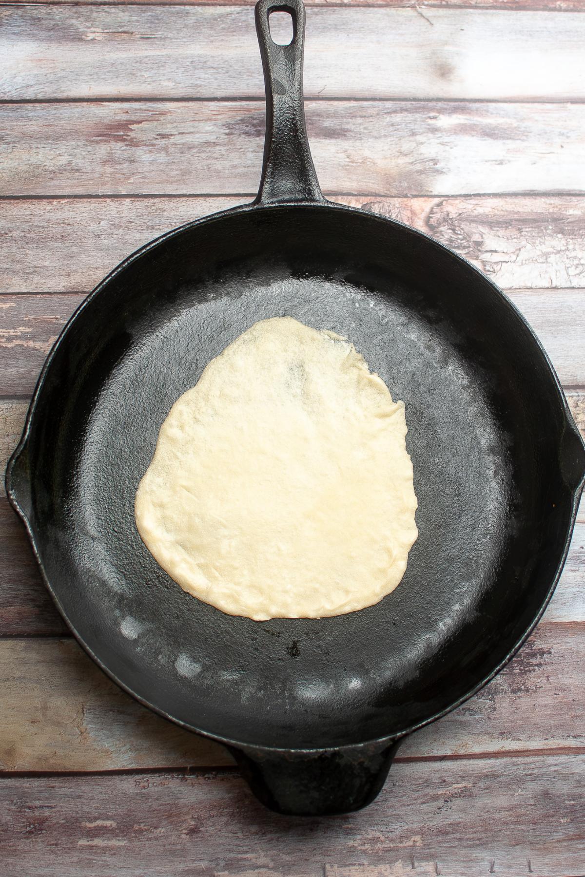 A piece of uncooked flatbread dough sits in the center of a black cast iron skillet on a wooden surface.