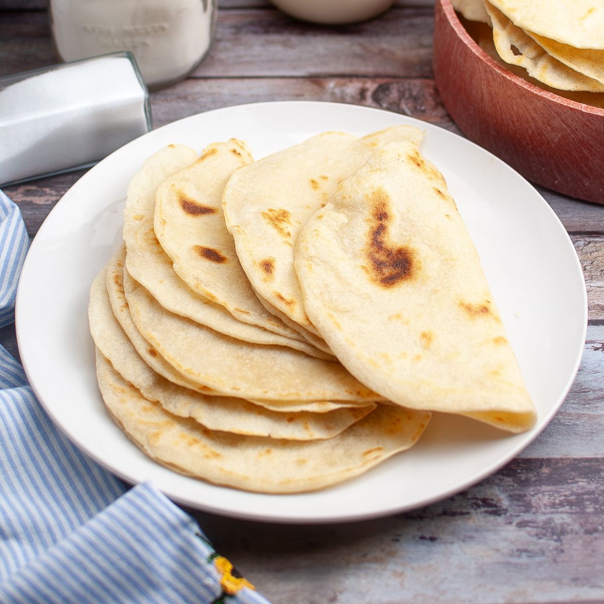 A white plate with several cooked flour tortillas stacked on it, displayed on a wooden surface next to a striped napkin and a wooden bowl.