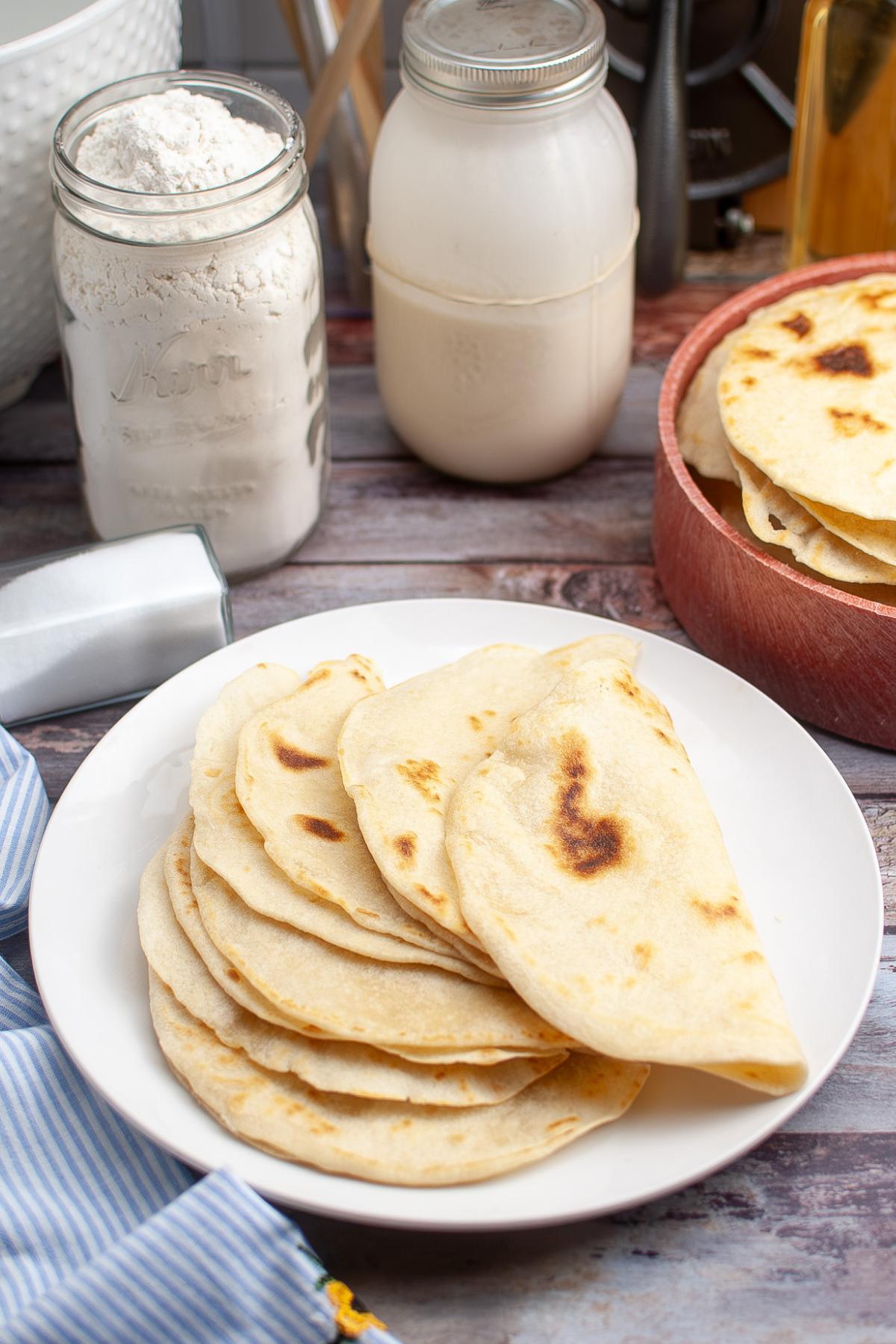 A plate of homemade flour tortillas sits on a table, with a bowl of more tortillas, a mason jar of flour, and a mason jar of liquid in the background.