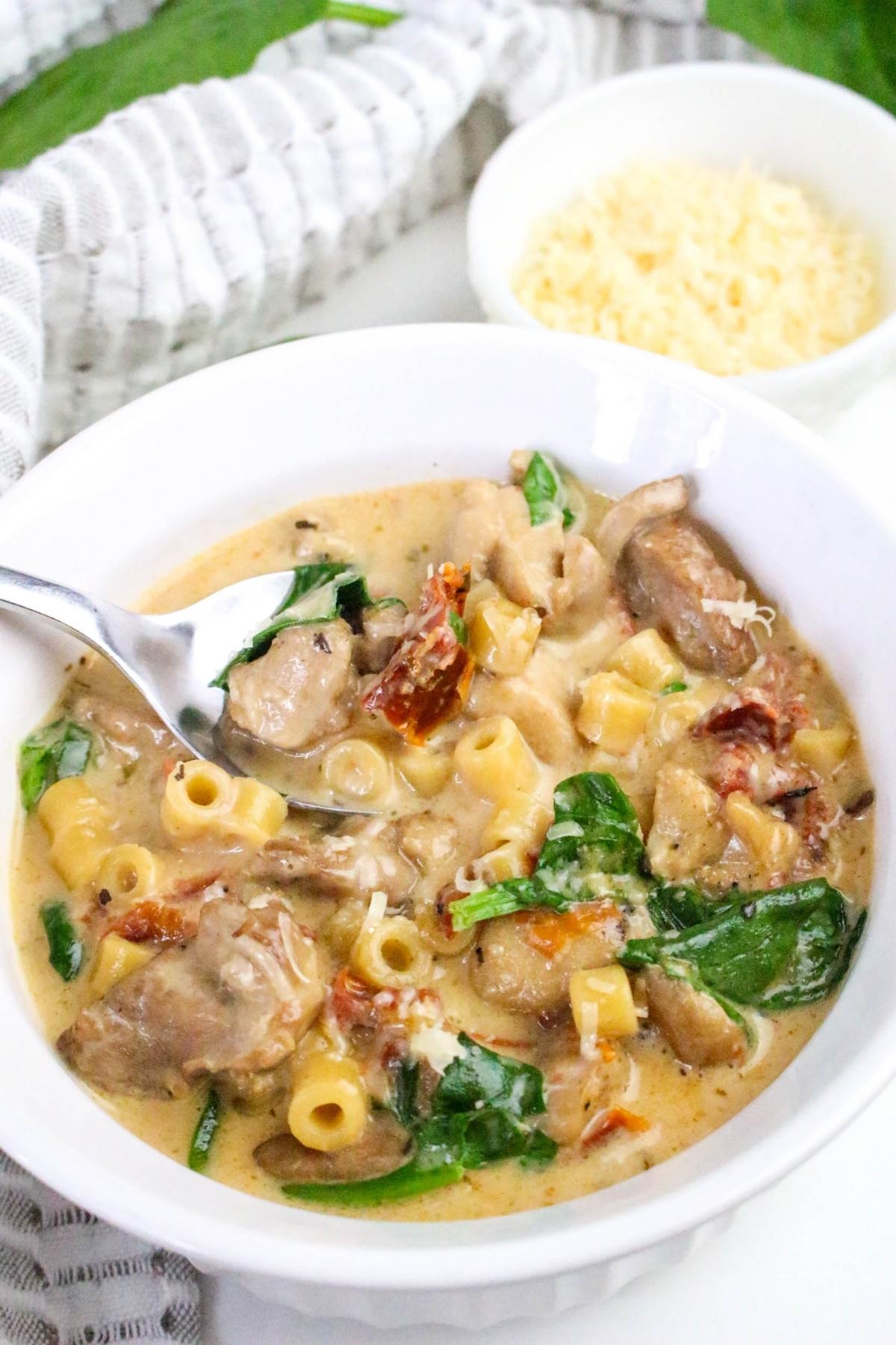 A white bowl of creamy pasta with mushrooms, sun-dried tomatoes, spinach, and grated cheese, with a spoon in the bowl and a small dish of cheese in the background.