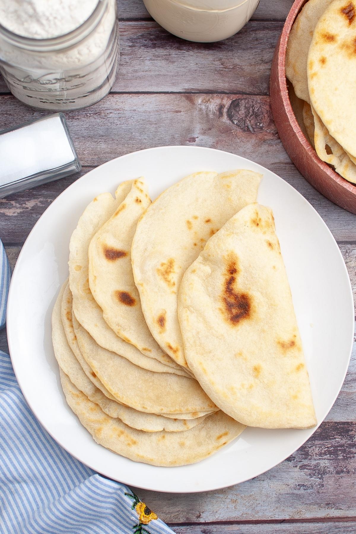A white plate with several folded flour tortillas on a wooden surface, with a cloth napkin and containers of flour and salt nearby.