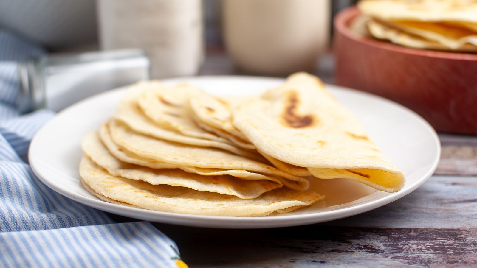 A white plate with several folded flour tortillas stacked on it, with a striped cloth and kitchen items in the background.