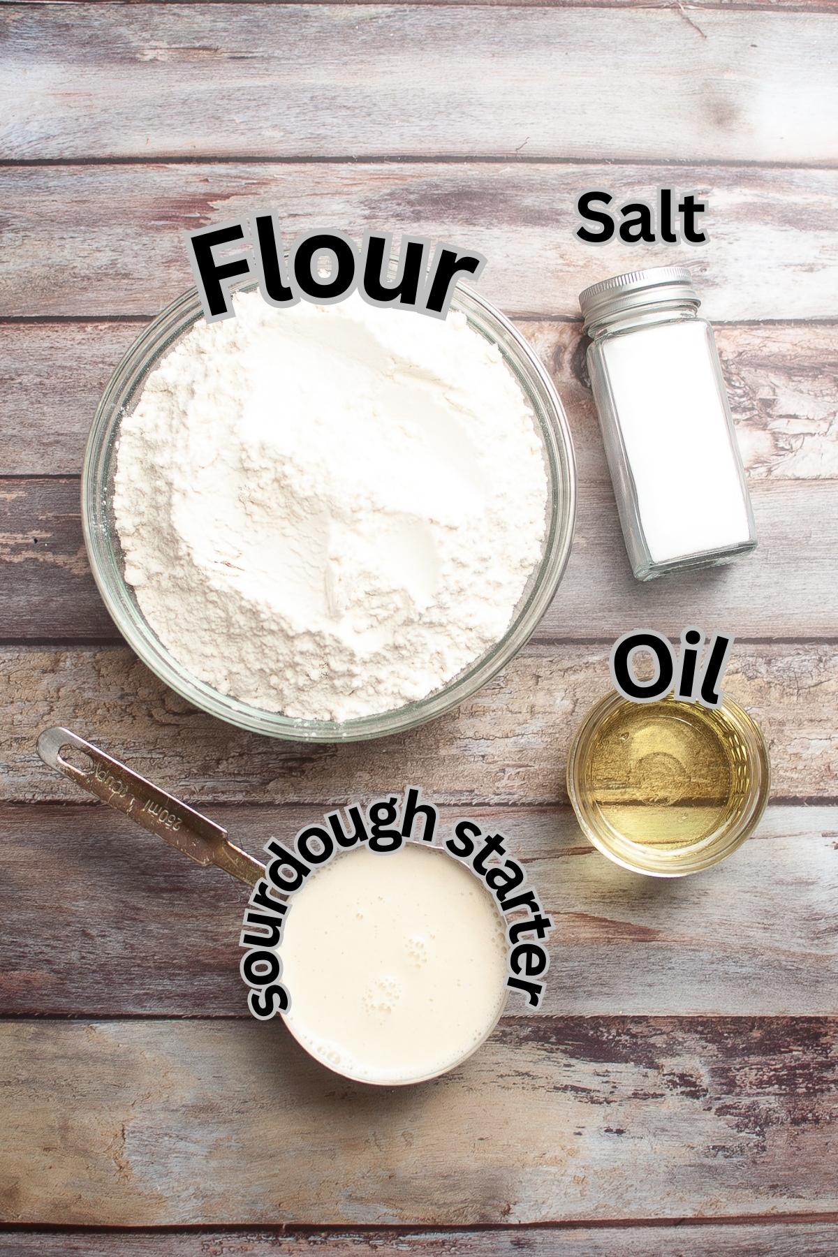 A bowl of flour, a glass jar of salt, a small cup of oil, and a measuring cup of sourdough starter are arranged on a wooden surface.