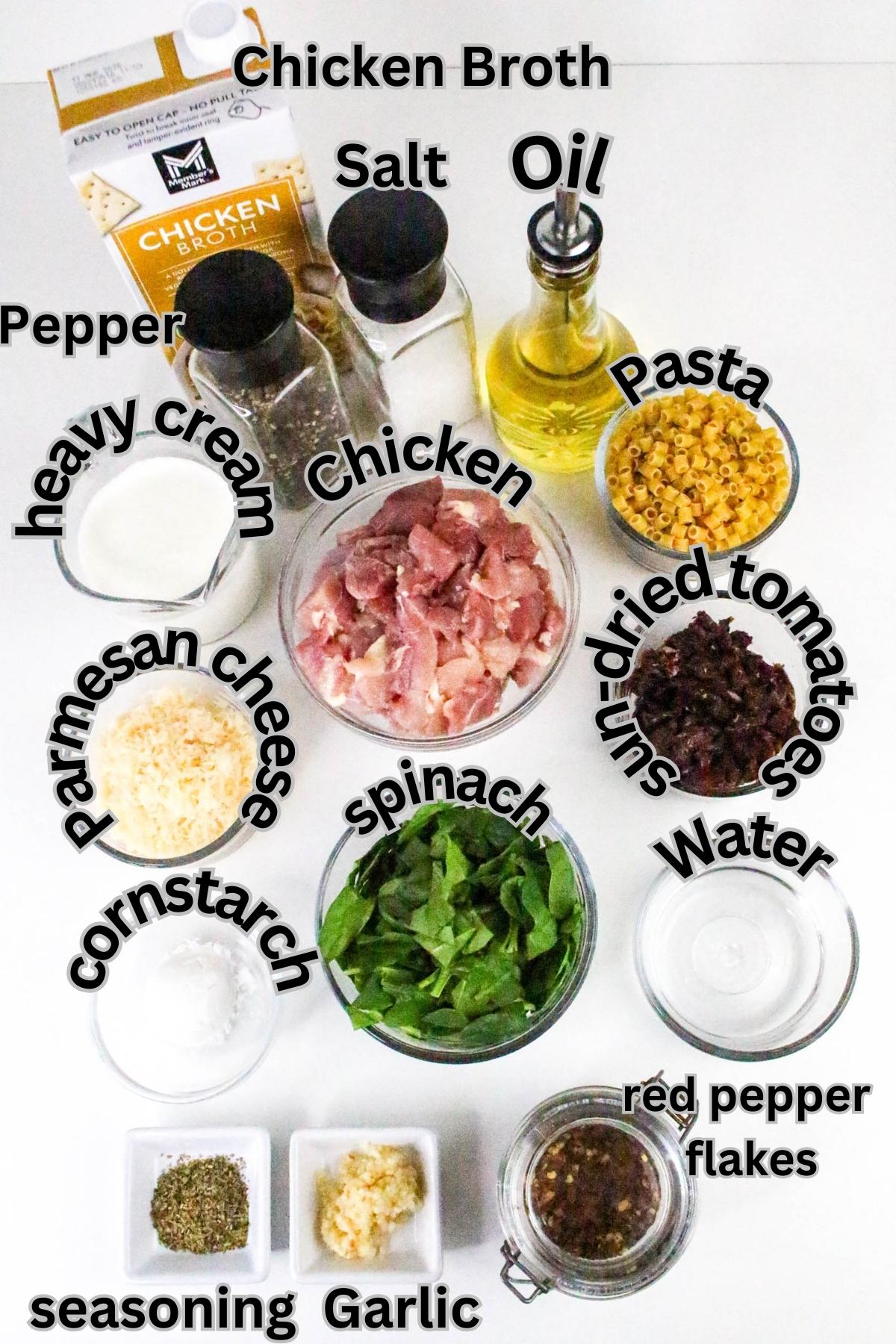 Ingredients for a pasta dish are arranged on a white surface, including chicken, spinach, dried tomatoes, pasta, broth, oil, garlic, spices, cream, parmesan, salt, pepper, and water.