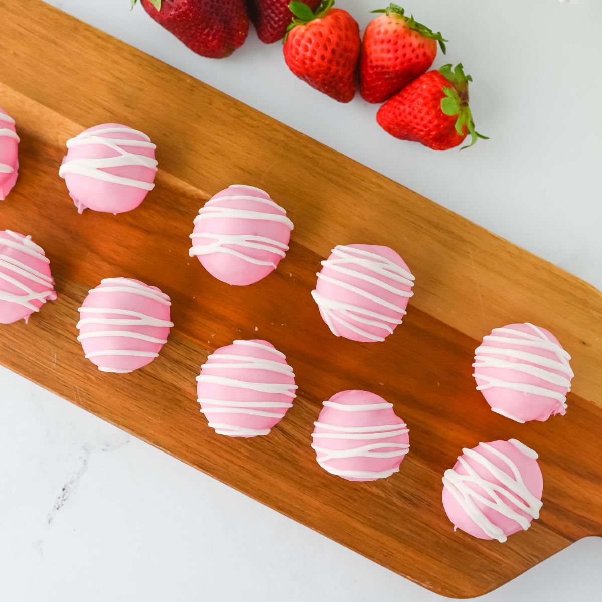 Pink strawberry-flavored cake balls with white drizzle on a wooden platter, with fresh strawberries in the background.