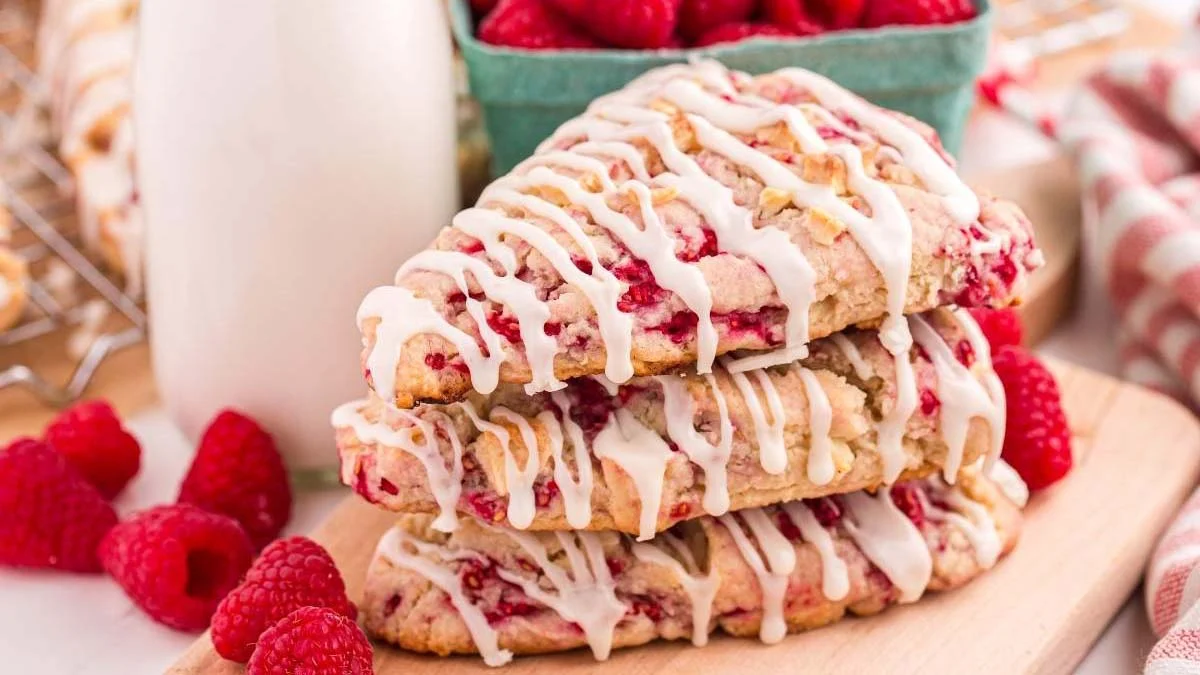 Stack of three White Chocolate Raspberry Scones drizzled with icing on a wooden board, surrounded by fresh raspberries and a bottle of milk.