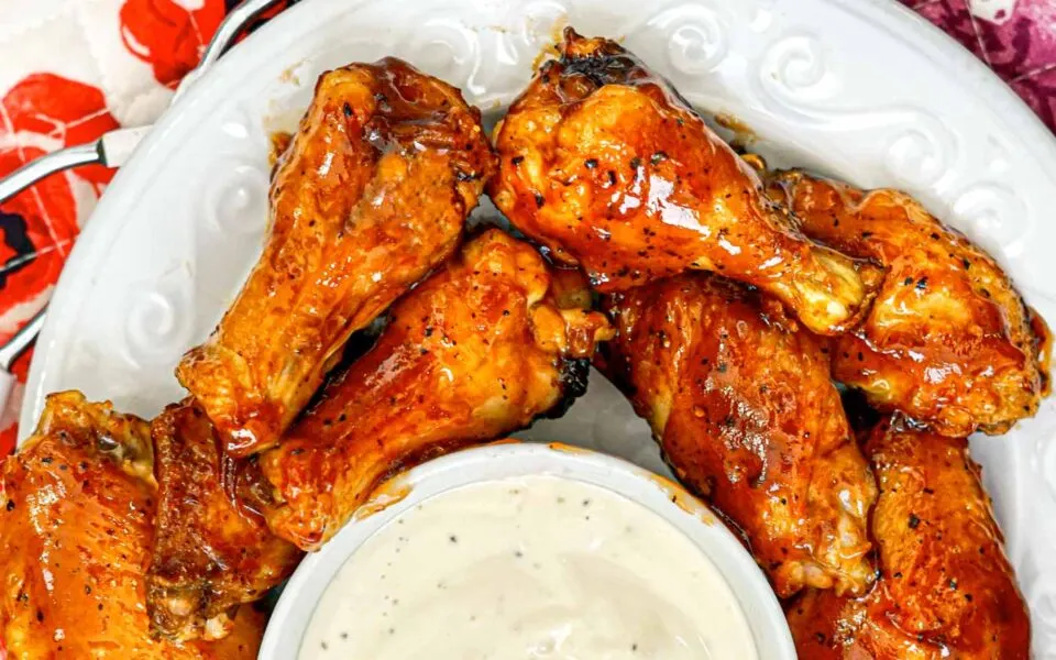 Chicken wings glistening with a rich, sticky barbecue sauce arranged on a white plate accompanied with a small bowl of creamy dipping sauce.