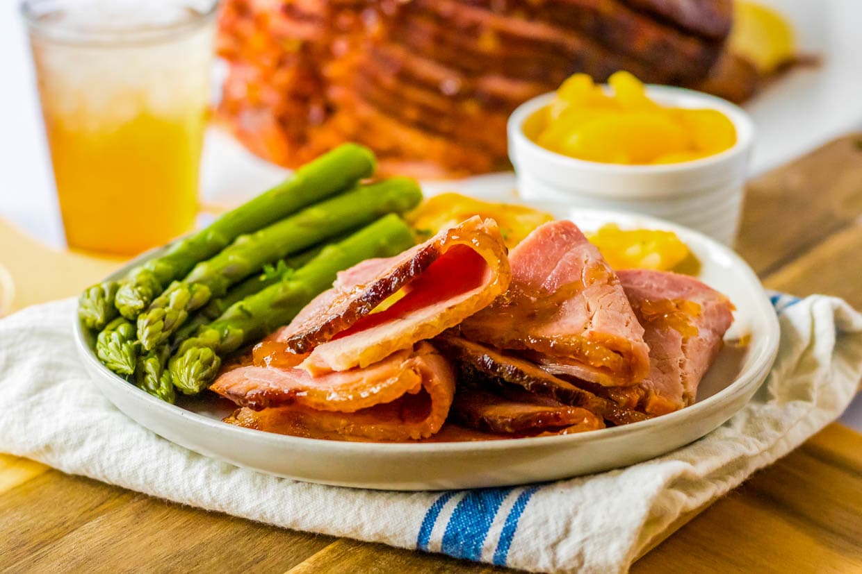 A plate with sliced glazed ham, cooked asparagus, and a side of peaches sits on a white napkin with a blue stripe—one of 23 Traditional Dinner Ideas perfect for a classic meal.