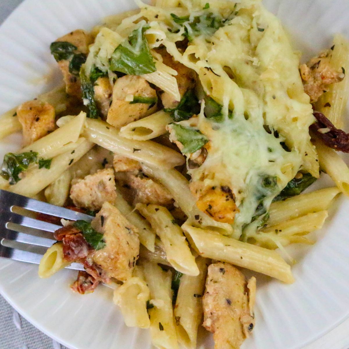 A plate of Marry Me Chicken Casserole features penne pasta with chicken, spinach, sun-dried tomatoes, and melted cheese, with a fork resting on the side.