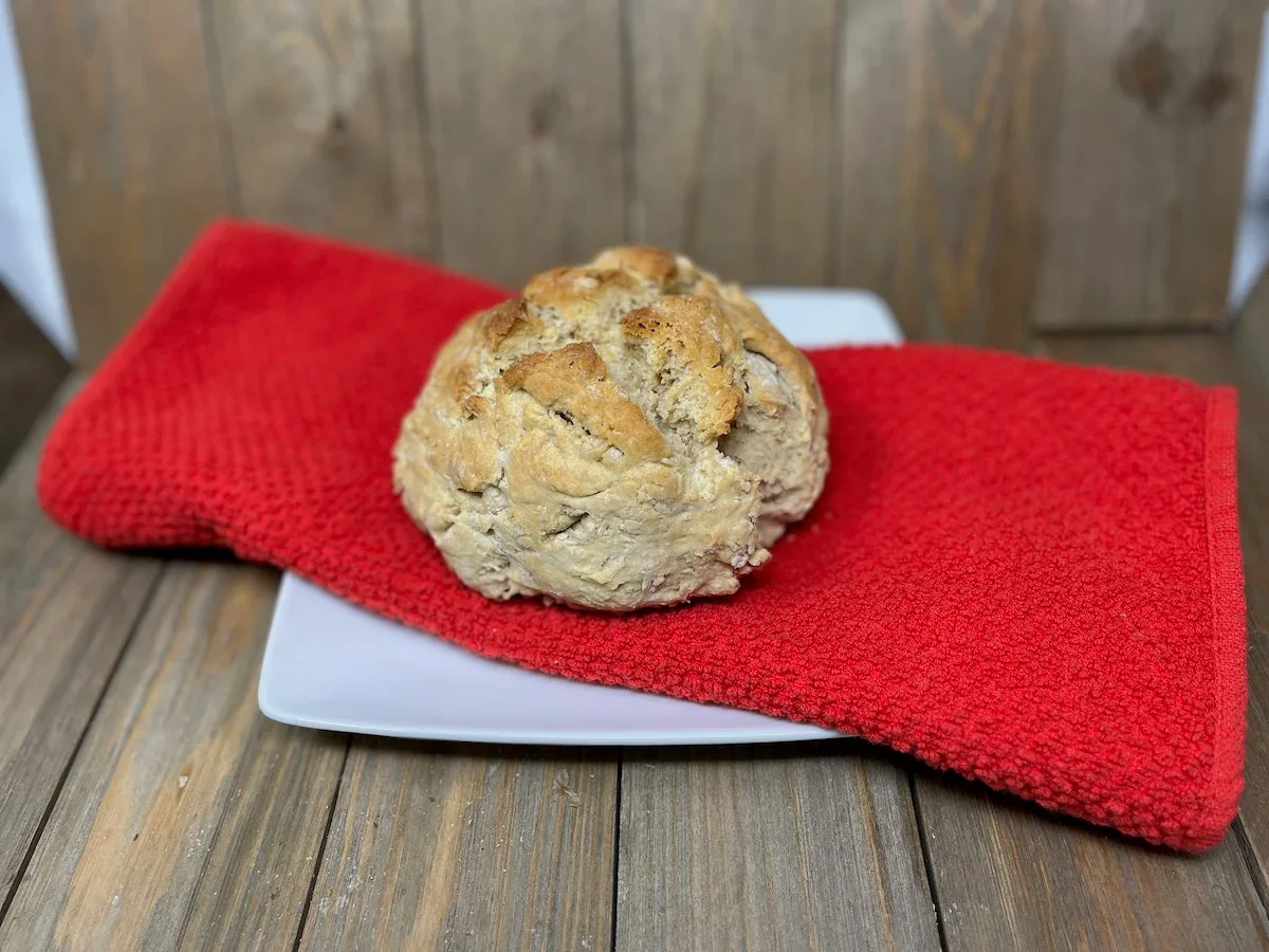 A round loaf of bread, inspired by our 14 easy and tasty bread recipes, sits on a white square plate with a red textured towel, placed on a wooden surface and background.