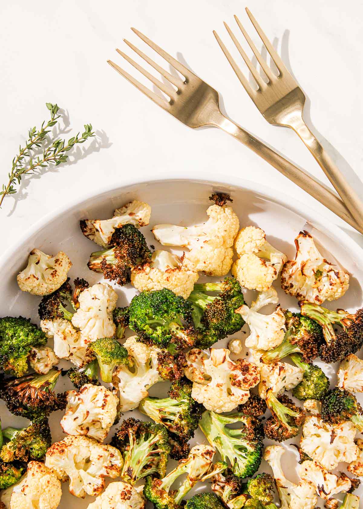 A white dish filled with roasted cauliflower and broccoli sits on a white surface beside two gold forks and a sprig of thyme.