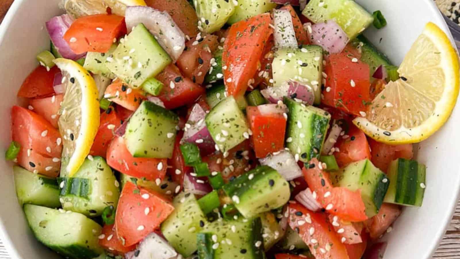 A bowl of salad with diced cucumbers, tomatoes, red onions, and lemon slices, topped with sesame seeds and herbs.