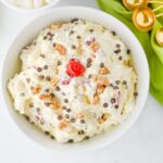 A white bowl filled with ambrosia salad topped with walnuts, mini chocolate chips, and a red cherry, placed next to a green napkin and gold measuring spoons.