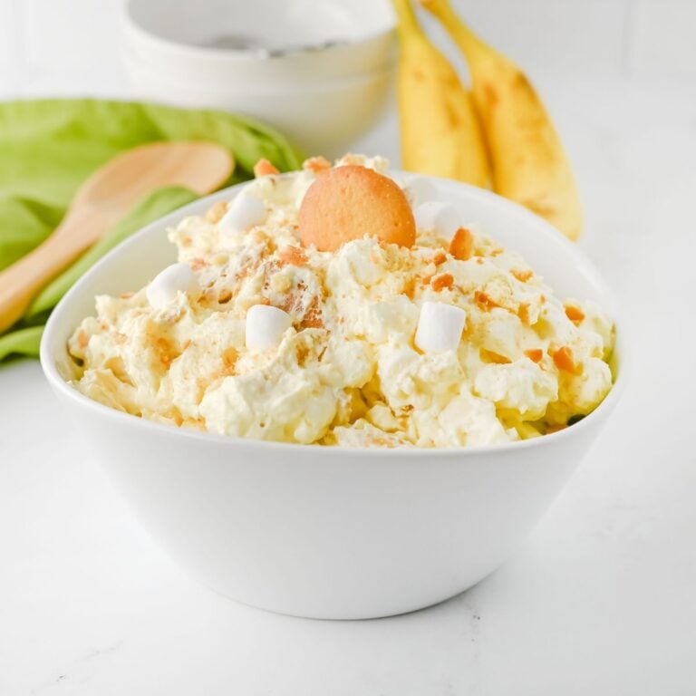 A white bowl filled with a creamy dessert topped with mini marshmallows, crumbled cookies, and a whole vanilla wafer; bananas and kitchen items in the background.