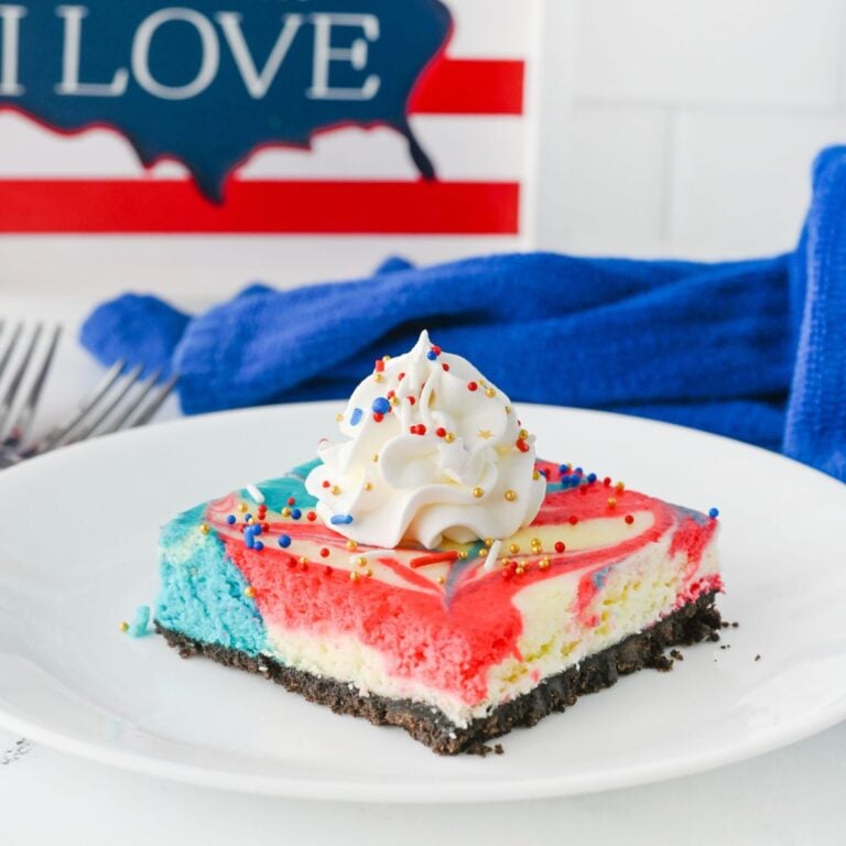 A square slice of red, white, and blue cheesecake with a chocolate crust, topped with whipped cream and sprinkles, sits on a white plate.