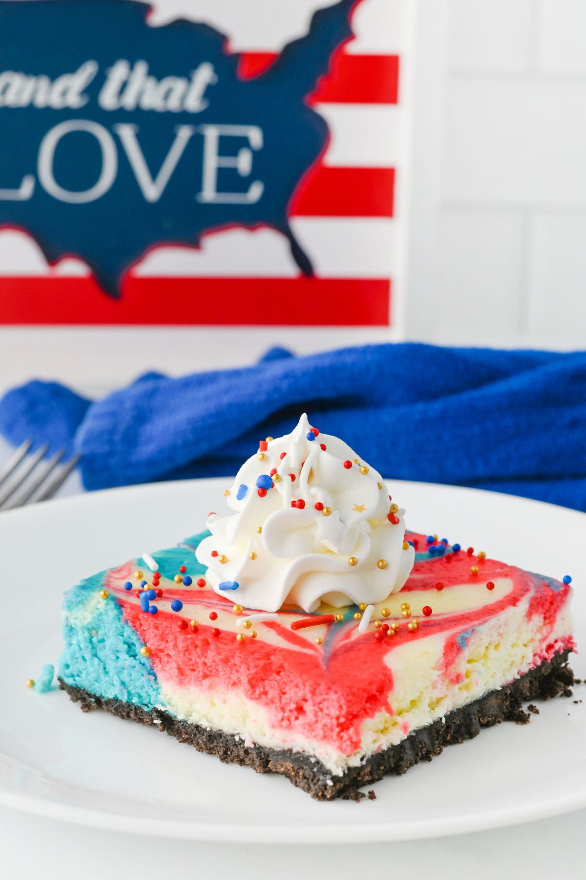 A slice of red, white, and blue marbled cheesecake with a cookie crust, topped with whipped cream and sprinkles, on a white plate. A patriotic sign is in the background.