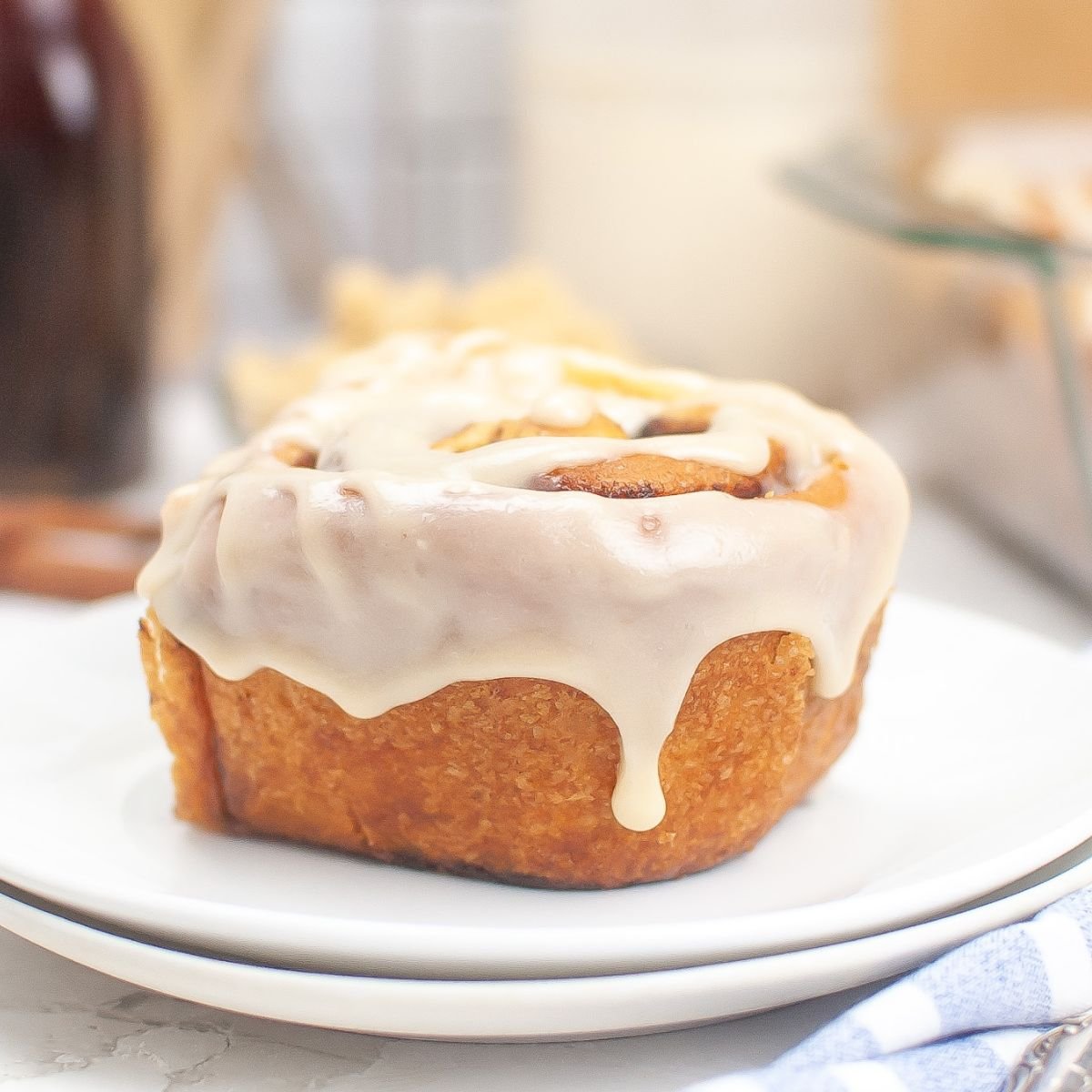 A cinnamon roll with white icing sits on a white plate, with a blurred background showing another plate and a light-colored napkin.