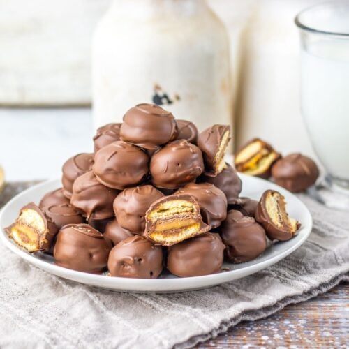 A plate piled with chocolate-covered sandwich cookies, some of which are cut open to show a layered filling, sits on a cloth napkin beside a glass of milk.