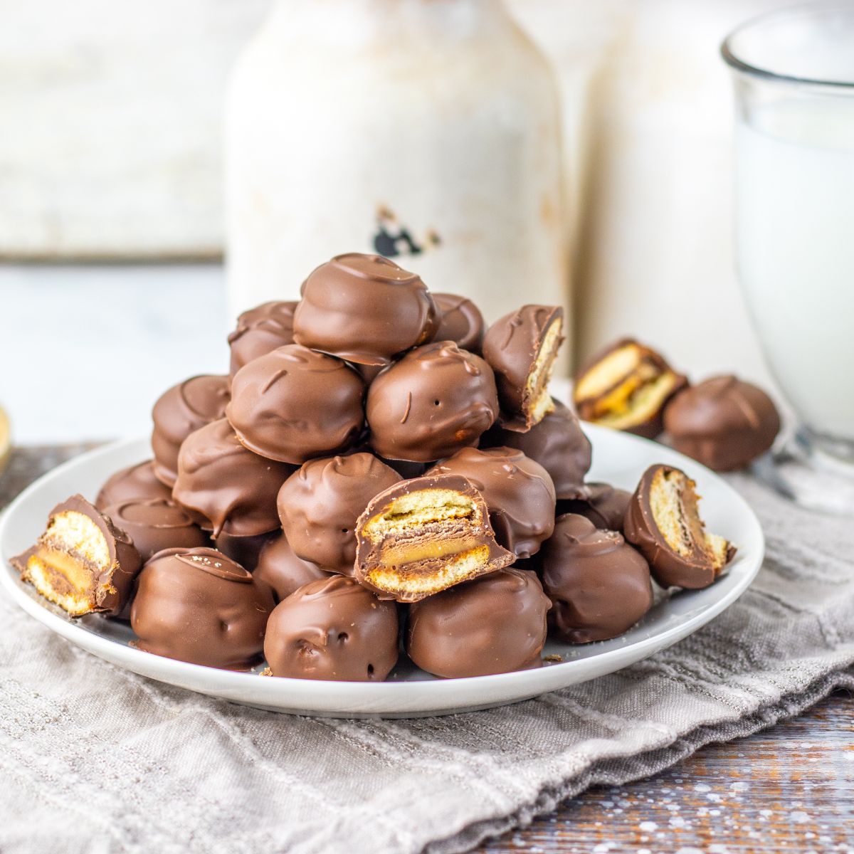 A plate piled with chocolate-covered sandwich cookies, some of which are cut open to show a layered filling, sits on a cloth napkin beside a glass of milk.