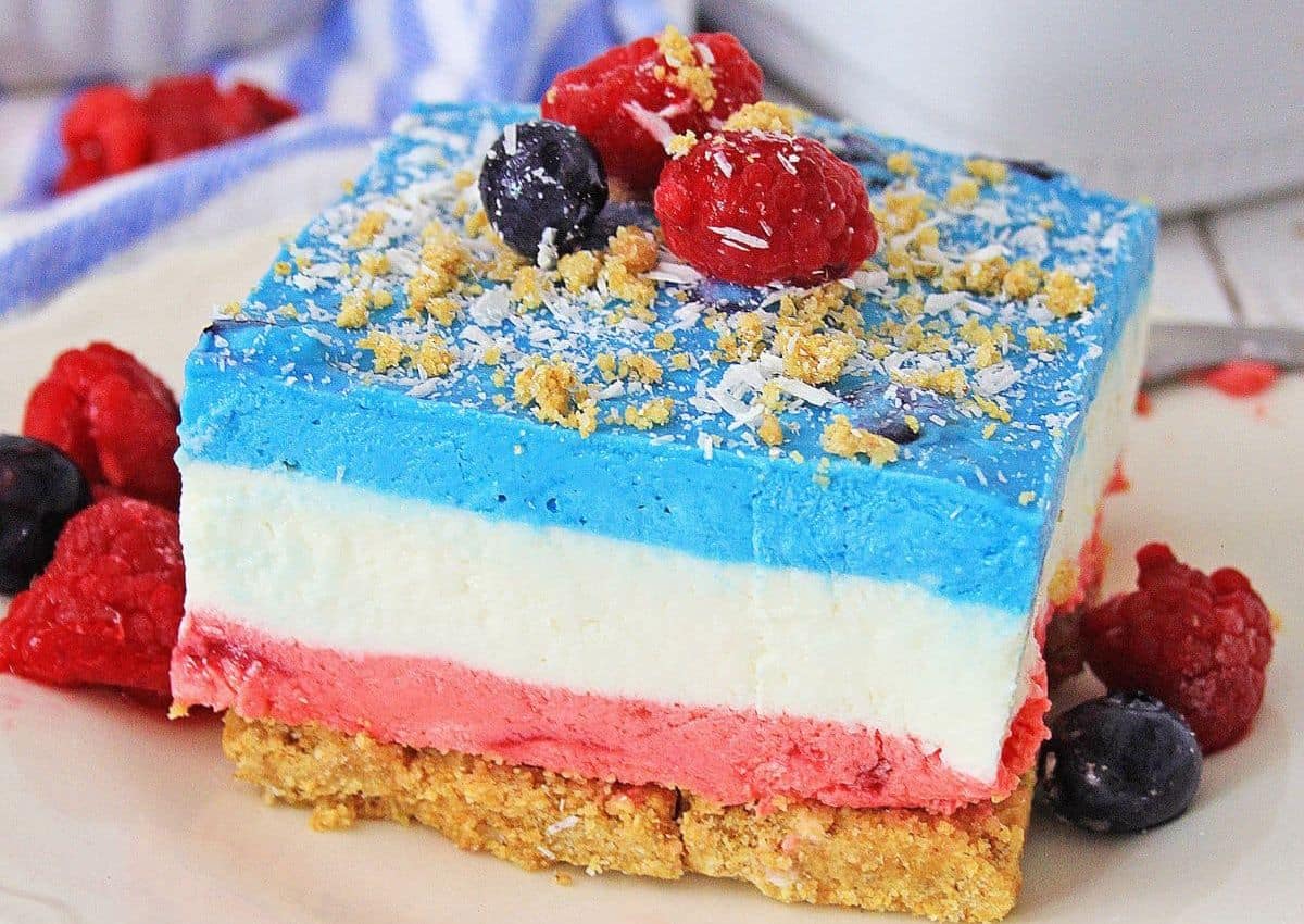 A slice of layered dessert with a crumb crust, red, white, and blue cream layers, topped with coconut flakes, crumbs, raspberries, and blueberries.