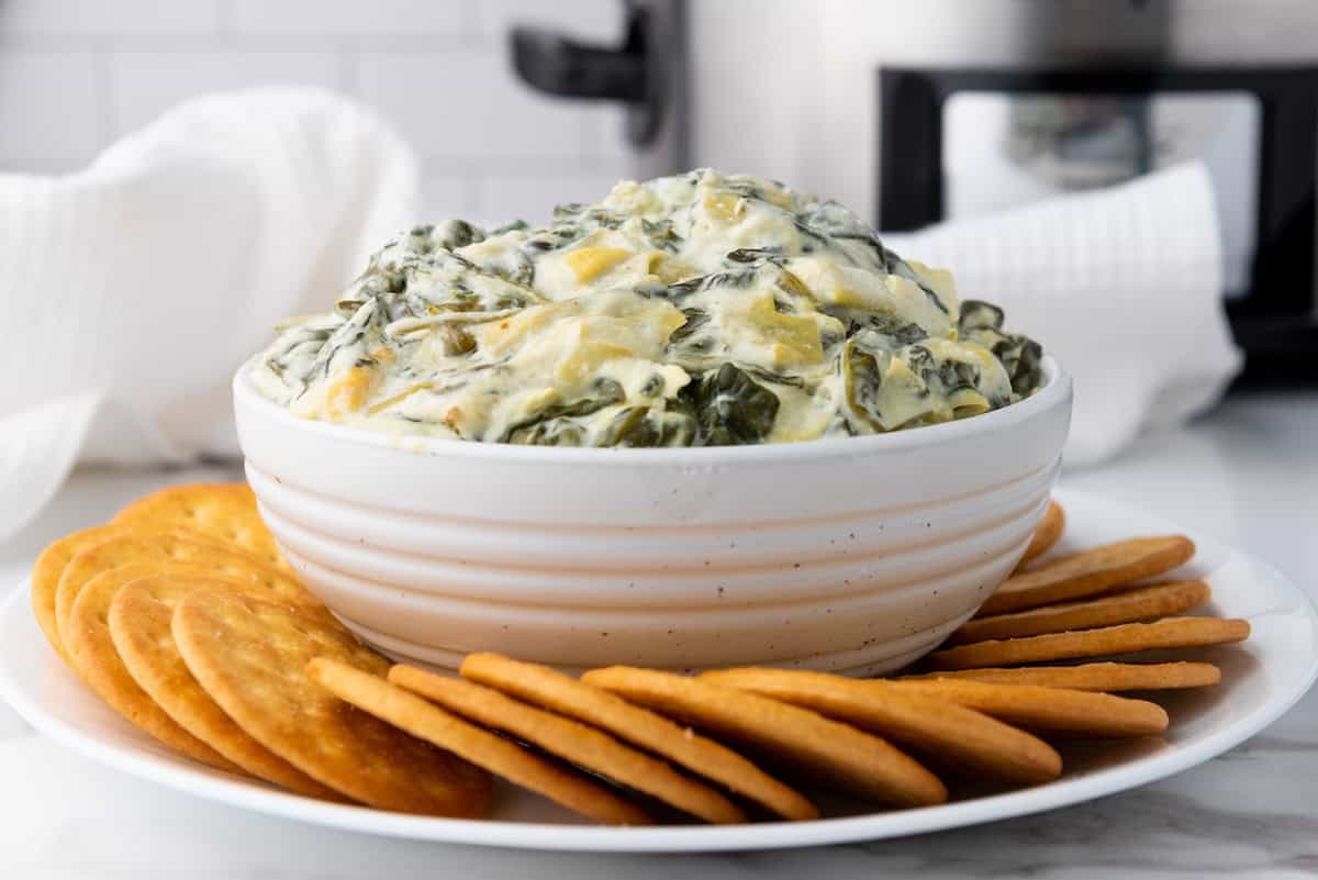 A bowl of creamy spinach artichoke dip surrounded by round crackers on a white plate.