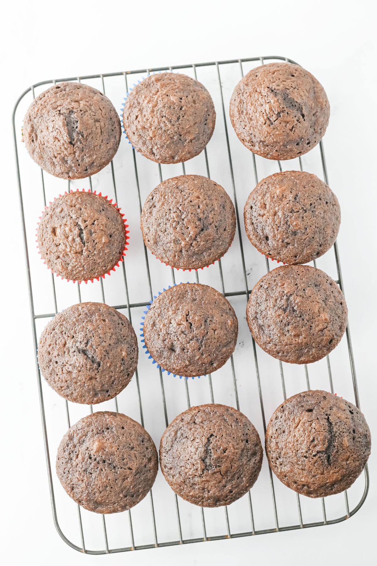 Twelve chocolate muffins in paper liners cooling on a metal wire rack against a white background.