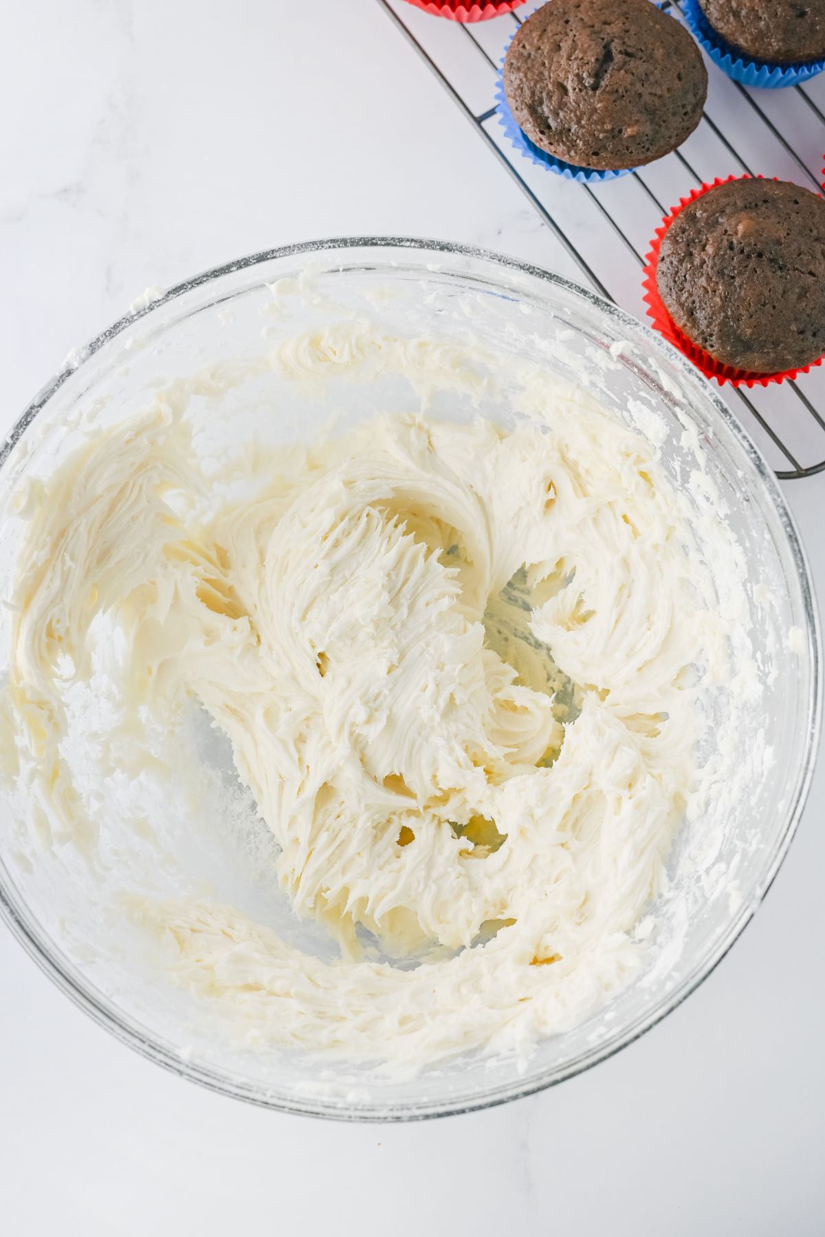 A glass bowl with whipped buttercream frosting sits on a white surface next to a cooling rack with chocolate cupcakes in blue and red liners.