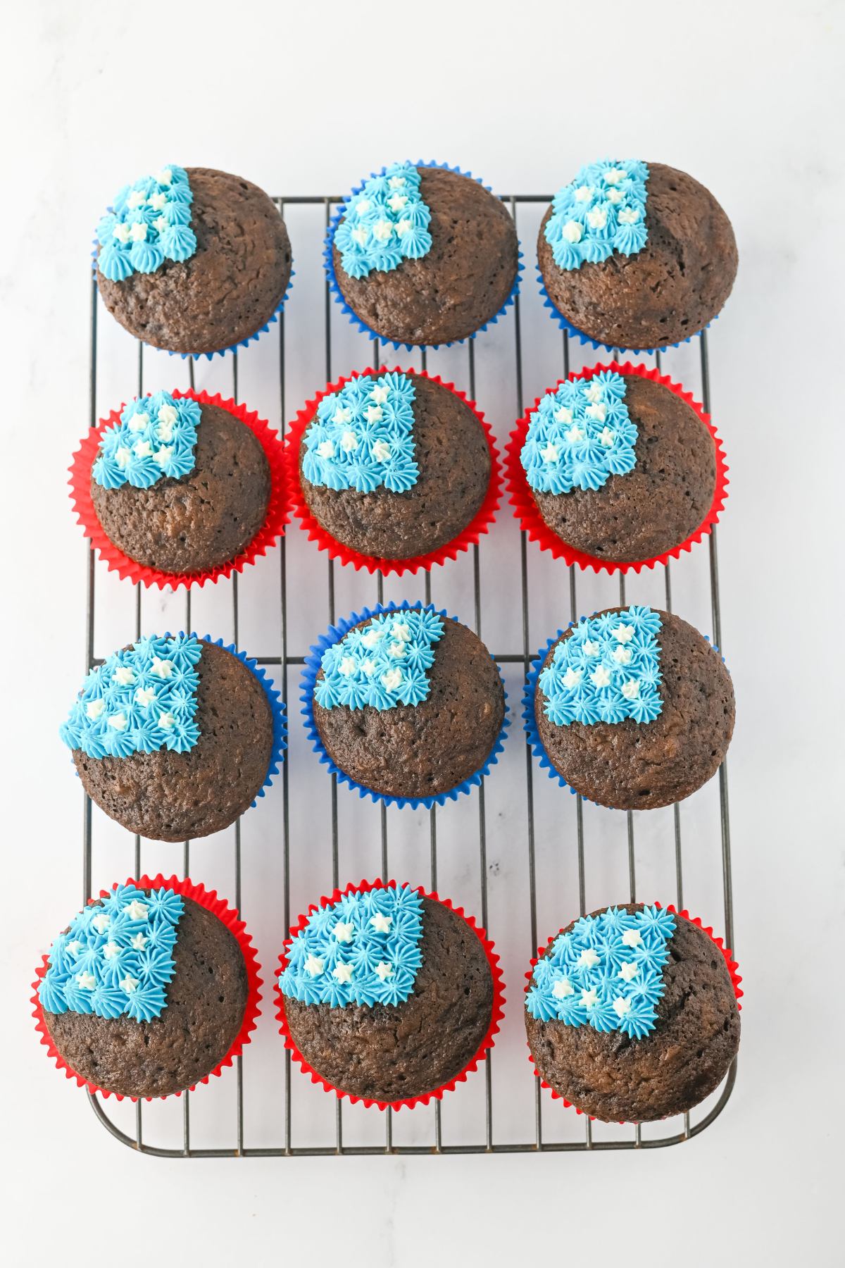 Twelve chocolate cupcakes in red and blue liners on a cooling rack, each topped with a square of blue star-shaped icing.
