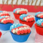 Chocolate cupcakes in blue and red liners topped with red, white, and blue piped frosting are arranged on a white surface with a decorative background.