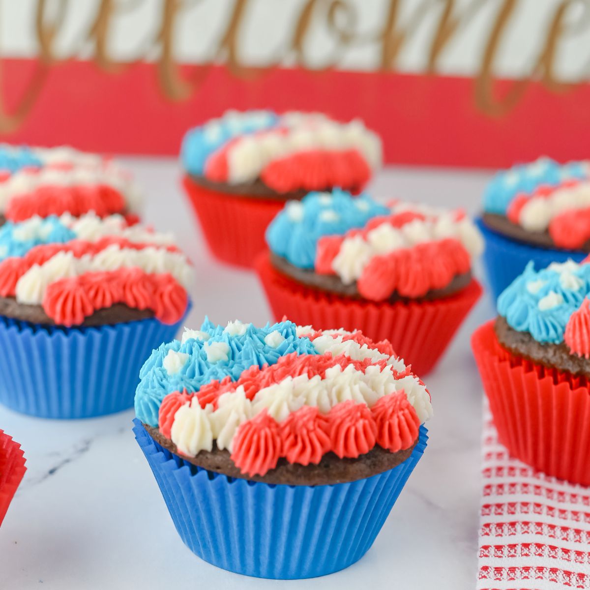 Chocolate cupcakes in blue and red liners topped with red, white, and blue piped frosting are arranged on a white surface with a decorative background.