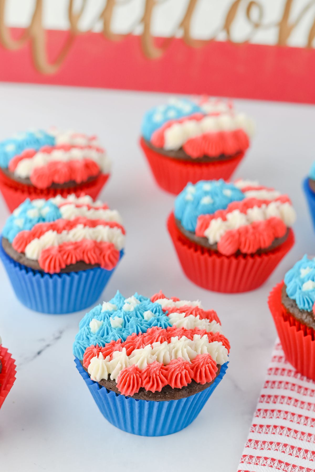Chocolate cupcakes in blue wrappers are decorated with red, white, and blue frosting in a striped pattern, arranged on a white surface.