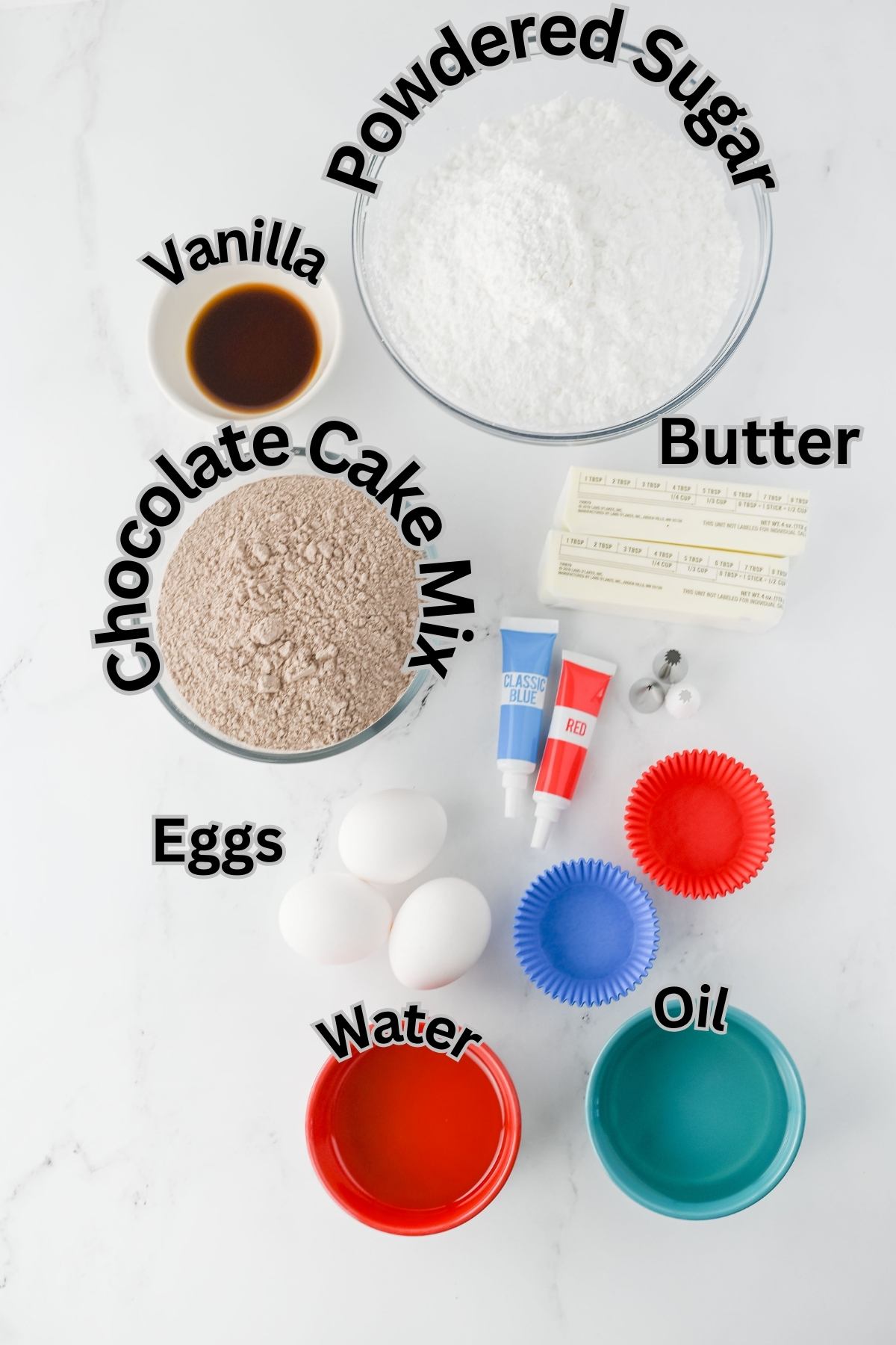 Baking ingredients including chocolate cake mix, powdered sugar, butter, eggs, vanilla, oil, water, measuring spoons, red and blue food coloring, and cupcake liners on a white surface.