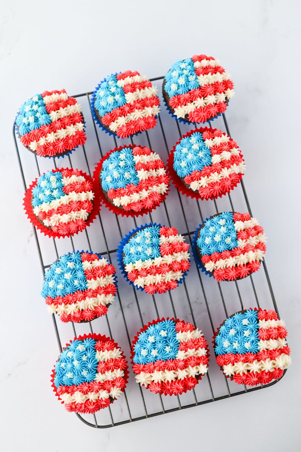 Cupcakes decorated with red, white, and blue frosting in the pattern of the American flag, arranged on a cooling rack.