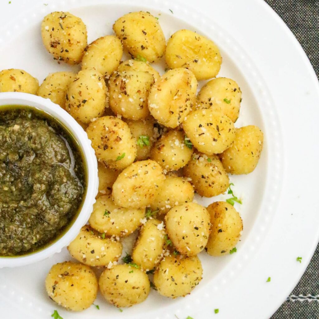A white plate with seasoned, baked gnocchi and a small bowl of green pesto sauce on the side.