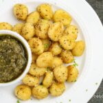 A white plate with seasoned, baked gnocchi and a small bowl of green pesto sauce on the side.