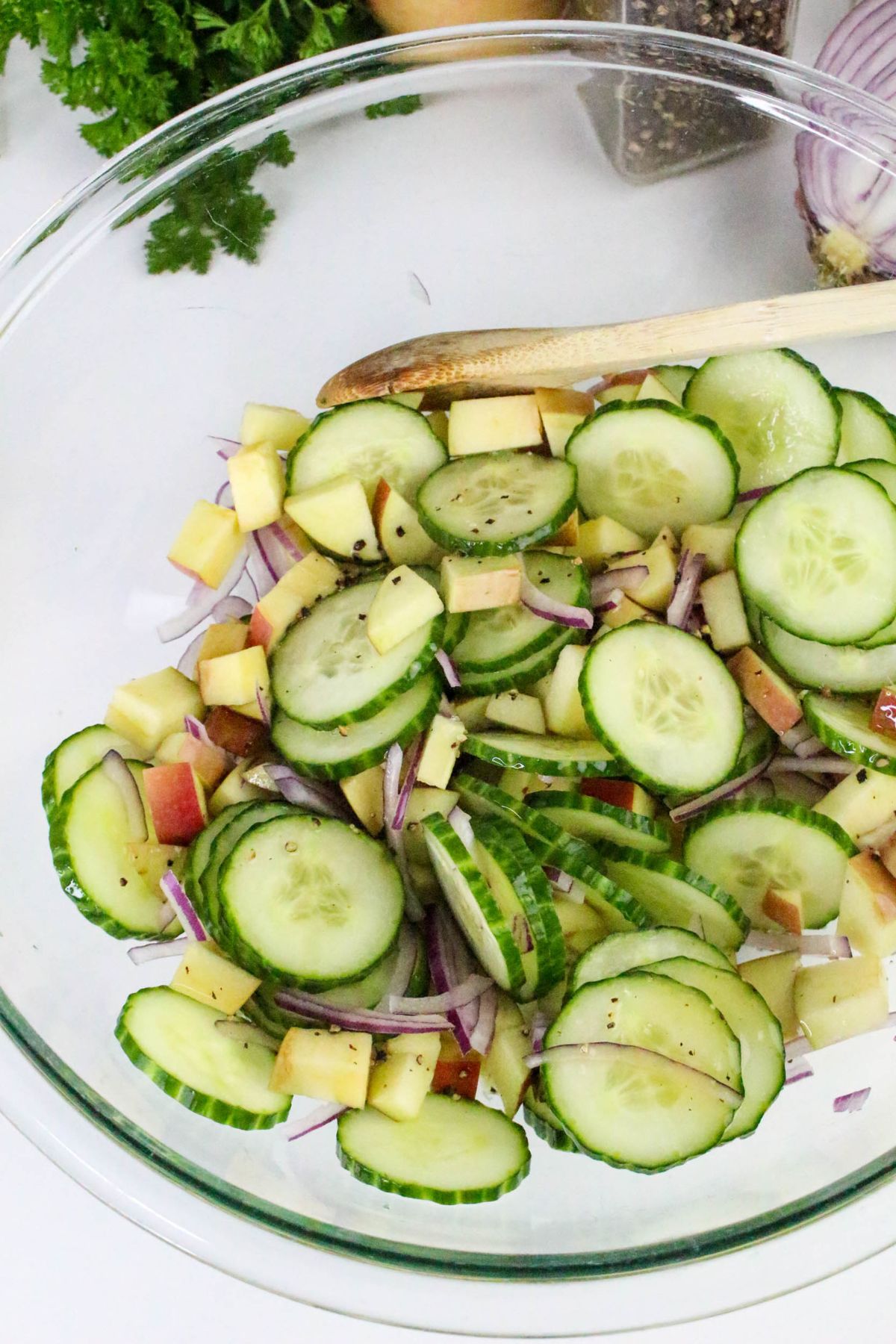 A glass bowl filled with sliced cucumbers, diced apples, and red onion, mixed with black pepper, next to a wooden spoon.